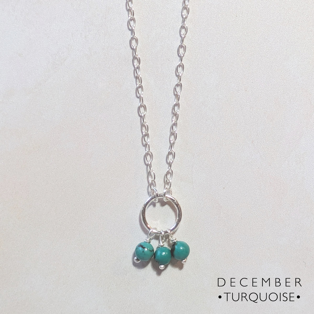 A silver chain birthstone necklace for December with a circular ring pendant and three small turquoise beads dangling from it, shown on a light background with the text 'DECEMBER • TURQUOISE •' in the lower right.