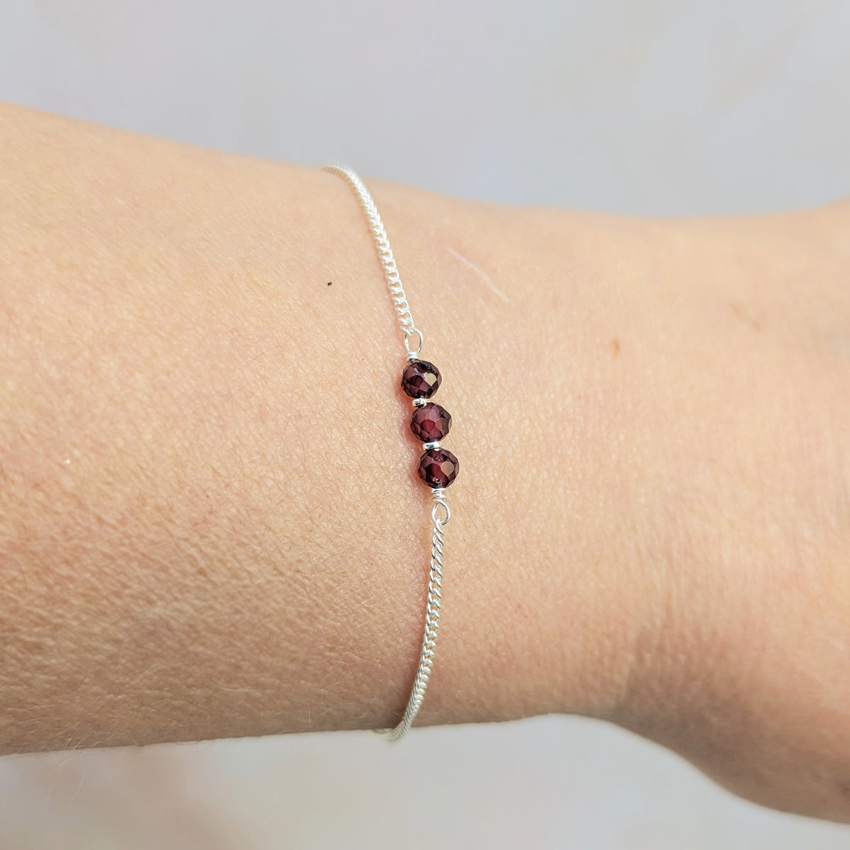 A delicate silver birthstone bar bracelet with three faceted dark red beads, shown worn on a real person's wrist.