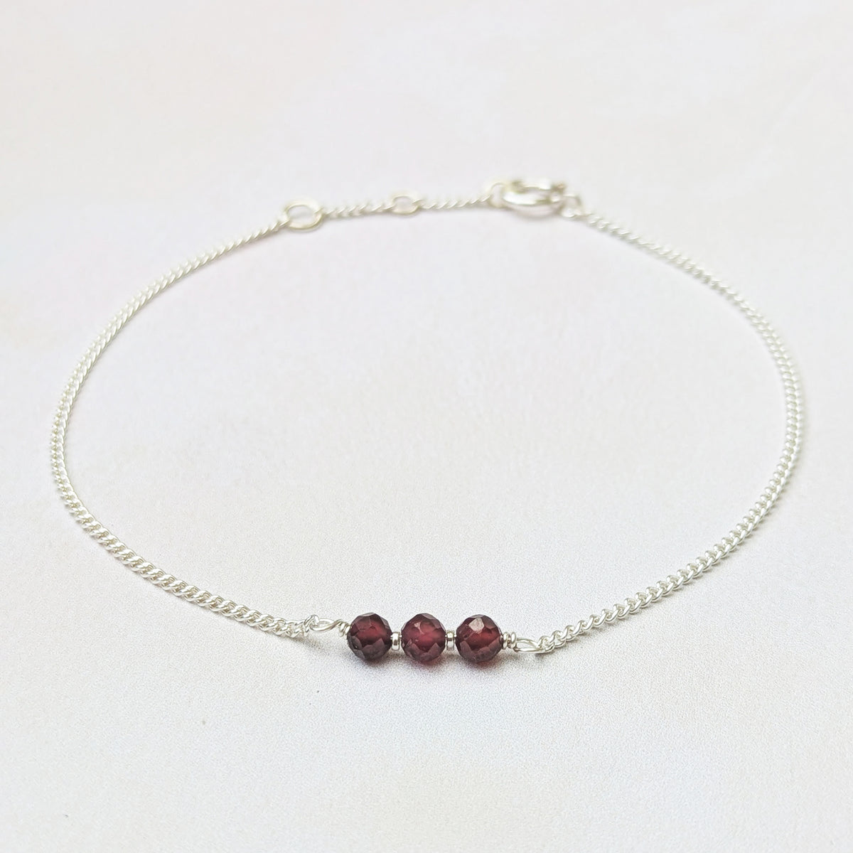 A delicate silver birthstone bar bracelet with three faceted dark red garnet beads at the centre, displayed on a clean white background.
