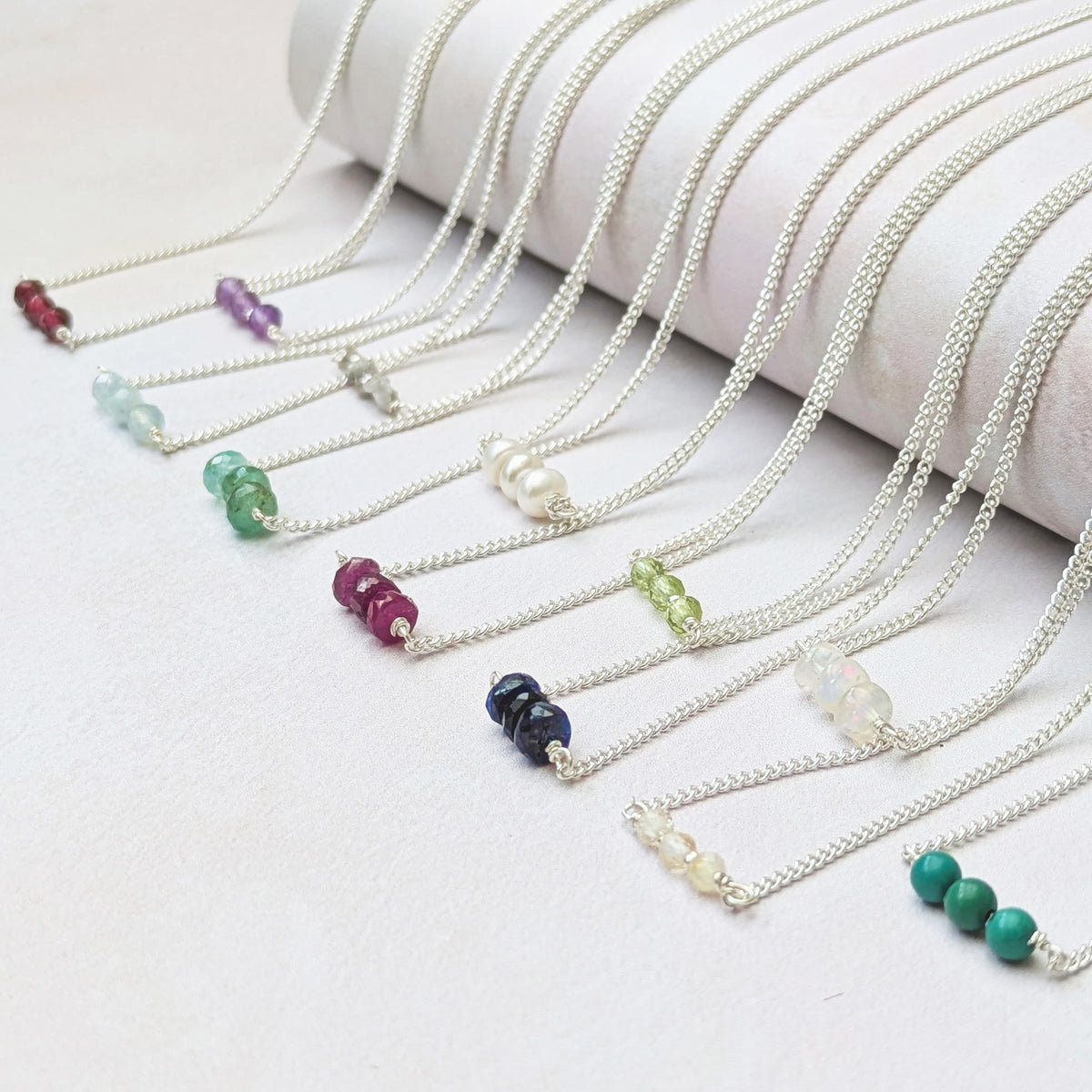 All 12 birthstone bar necklaces with colourful natural gemstone beads, arranged diagonally on a white surface next to a white cylindrical display stand.