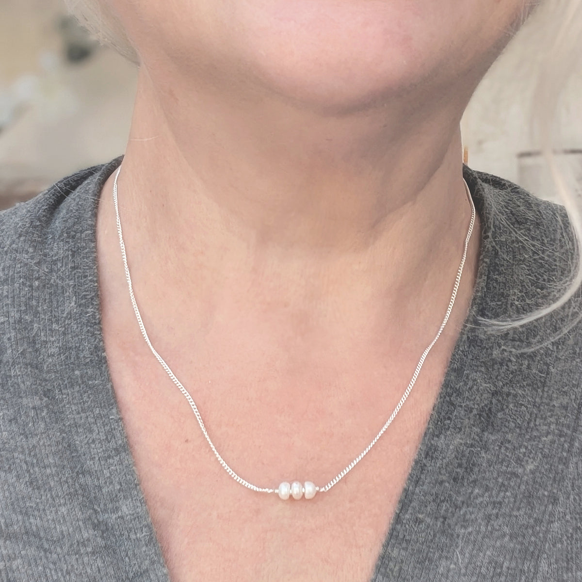 A close-up of a person wearing a delicate silver chain necklace with three small white pearls at the center, styled with a gray V-neck top.