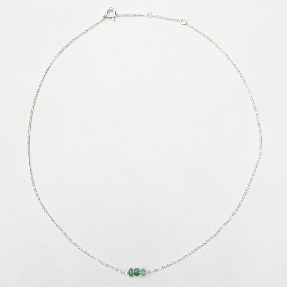A delicate silver chain necklace with three small faceted Emerald beads at the centre, displayed on a clean white background.