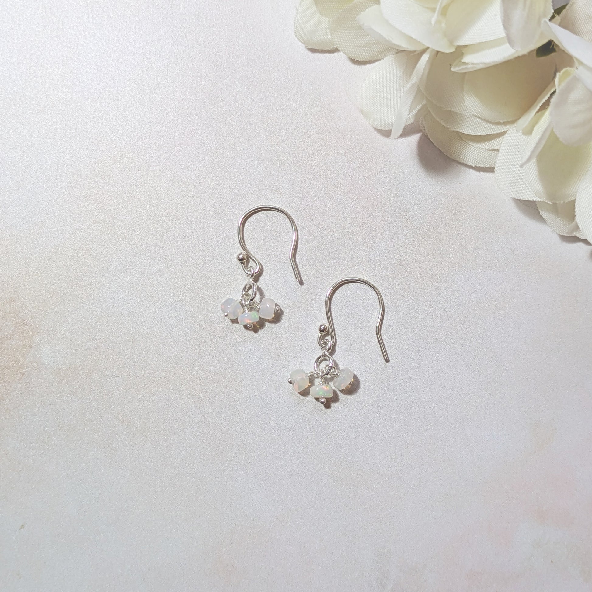 Silver dangling earrings with Opal gemstones on a neutral background with white flowers.