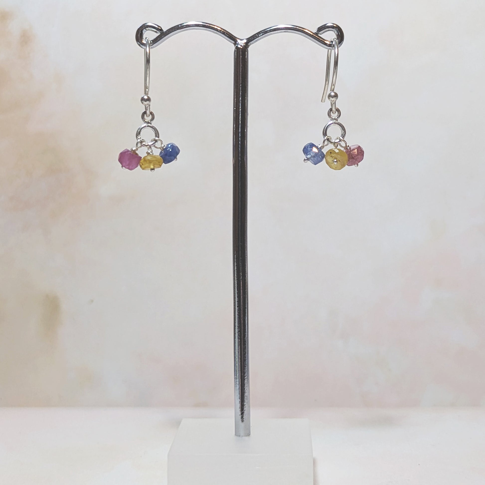 Pair of sterling silver earrings with pink, blue and yellow Sapphire beads on a stand against a light background.