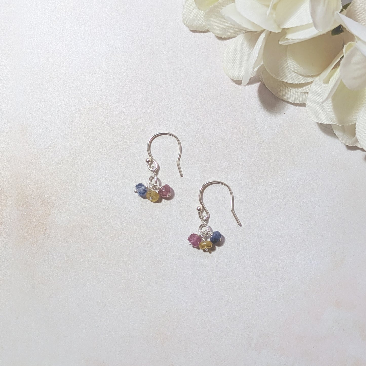 Silver earrings with blue, pink and yellow sapphire beads on a pale neutral background with white flowers.  