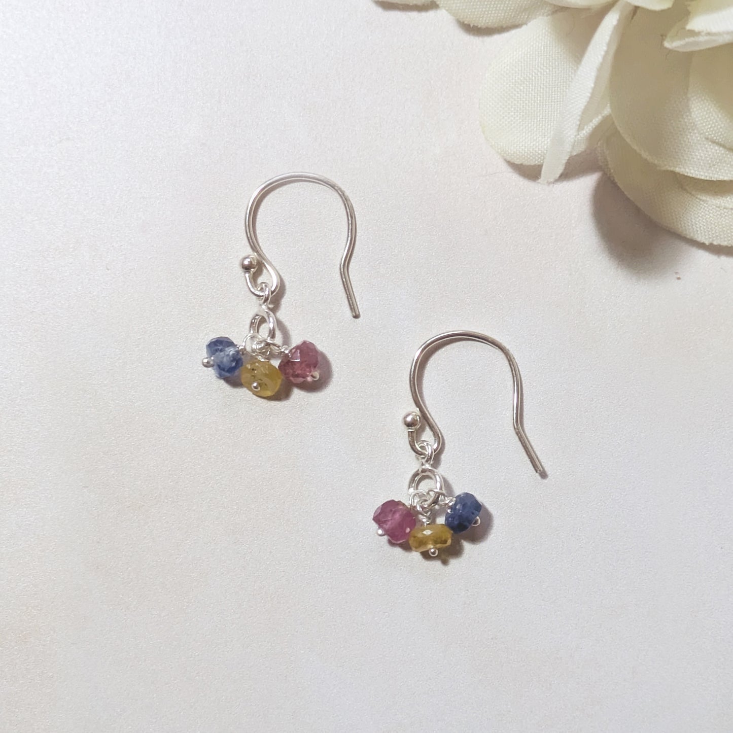 Silver drop earrings with multicoloured Sapphire gemstone beads on a pale surface with white flowers in the background.