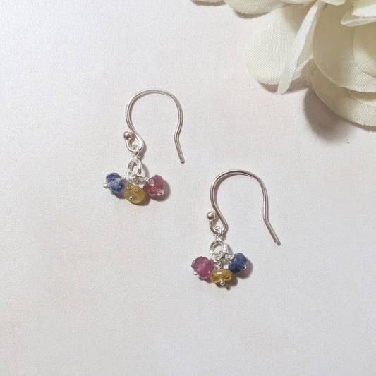 Silver drop earrings with multicoloured Sapphire gemstone beads on a pale surface with white flowers in the background.