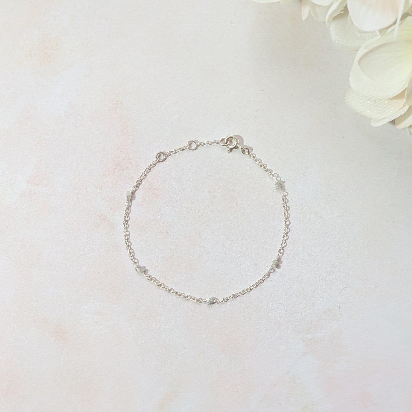 Silver bracelet with Diamond nugget beads on a blush pink surface with white flowers in the corner.