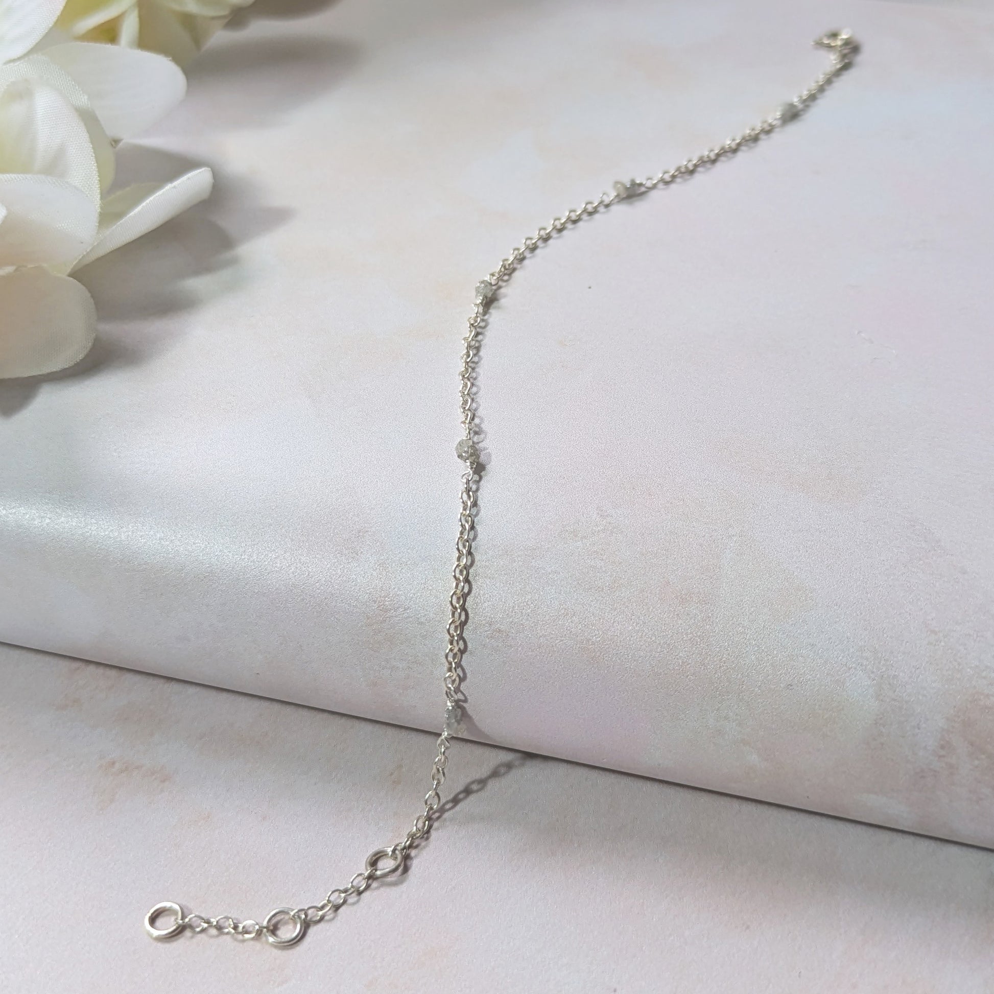 Silver station bracelet with rough Diamond nugget beads on a light surface with white flowers in the background.