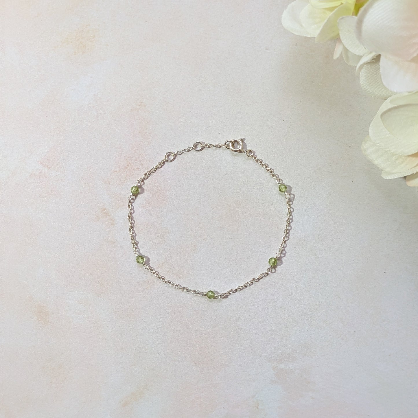 Silver stationed chain bracelet with Peridot gemstone beads on a light marble surface with flowers.
