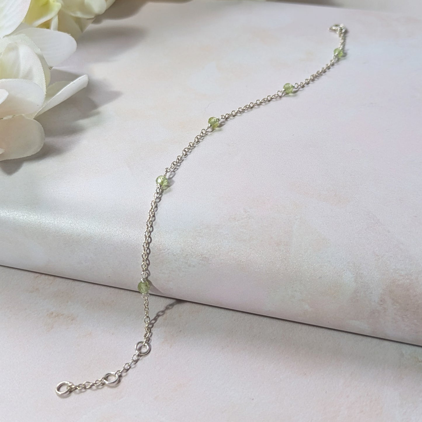 Silver station bracelet with green Peridot gemstone beads on a white surface with flowers in the background.
