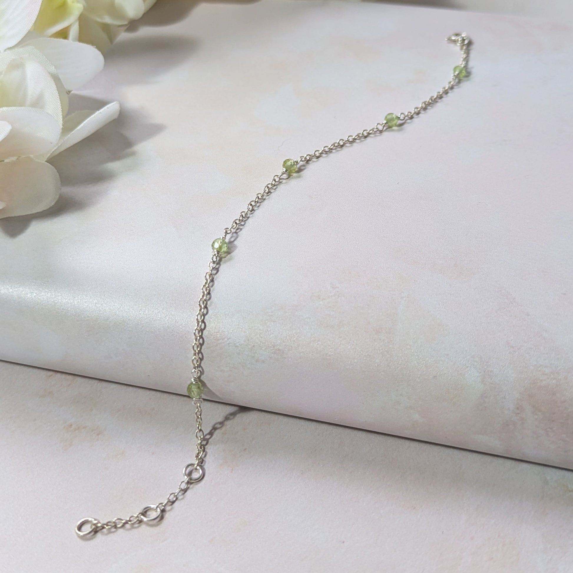 Silver station bracelet with green Peridot gemstone beads on a white surface with flowers in the background.