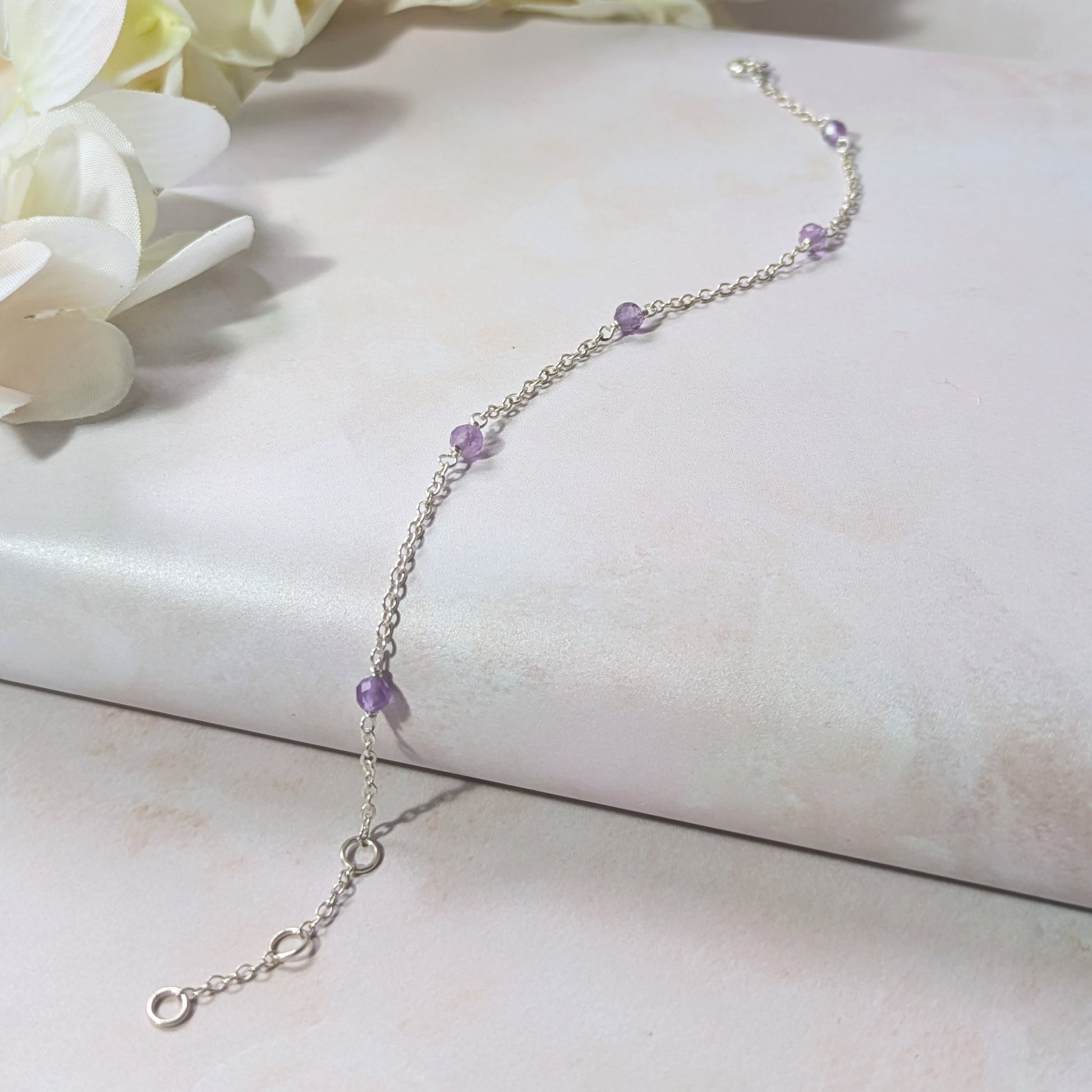 Silver station bracelet with Amethyst gemstone beads on a white surface with flowers in the background.