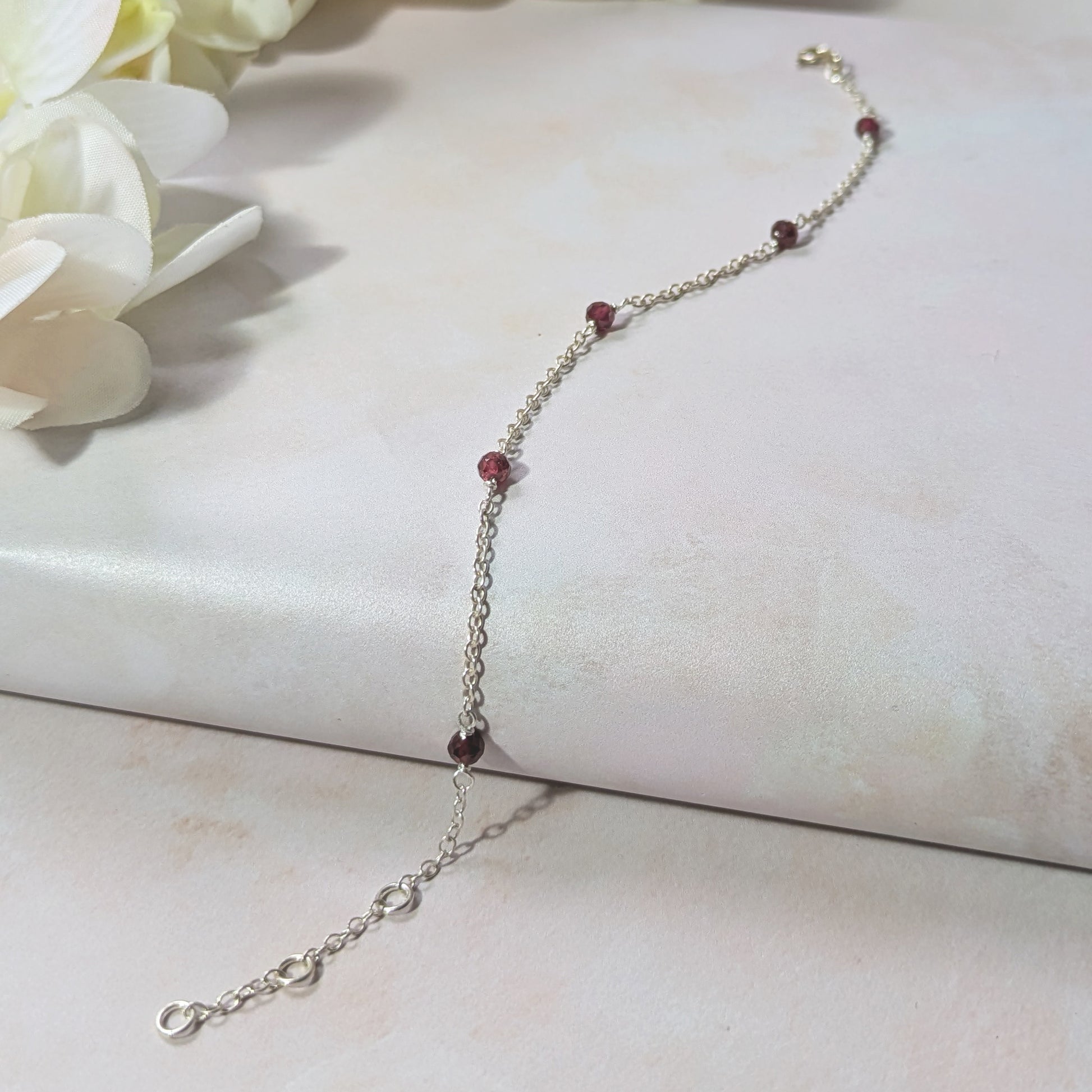 Silver stationed chain bracelet with red Garnet gemstone beads on a white surface with flowers.