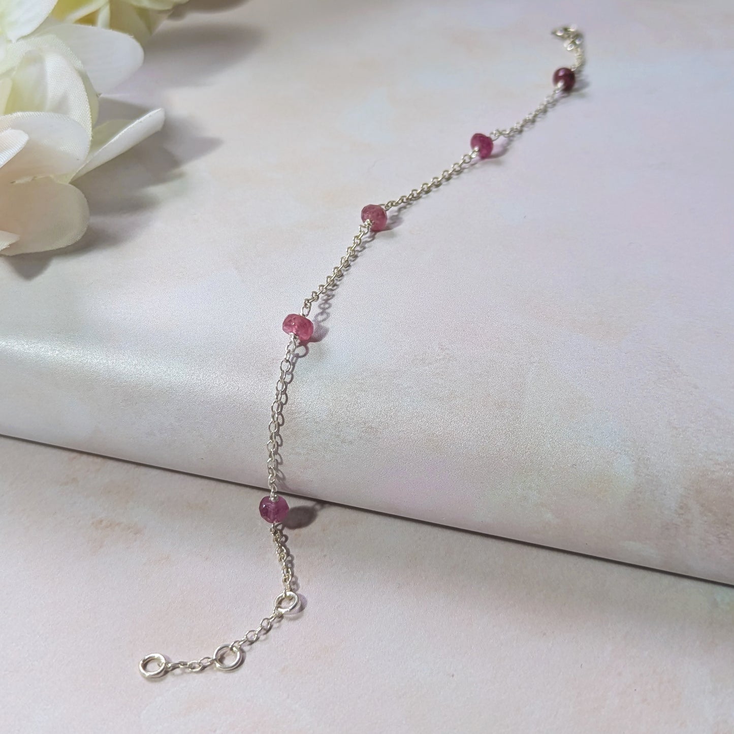 Silver bracelet with graduated Ruby gemstone beads on a light surface with flowers in the background.
