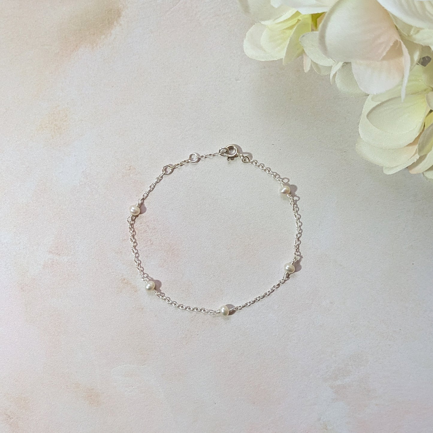 Silver station bracelet with freshwater pearls on a light marble surface with flowers in the background.