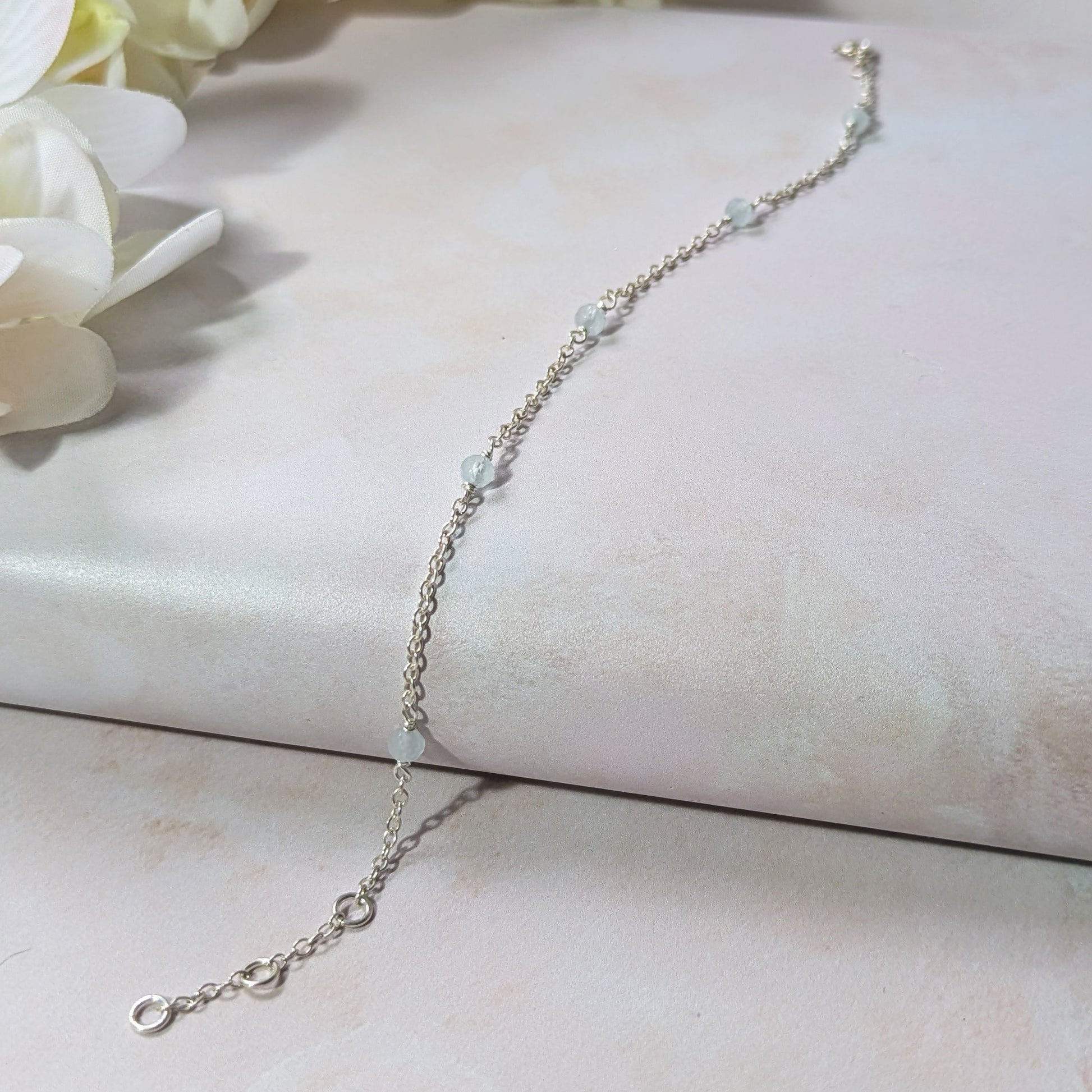Silver stationed bracelet with Aquamarine gemstone beads on a light pink surface with white flowers.