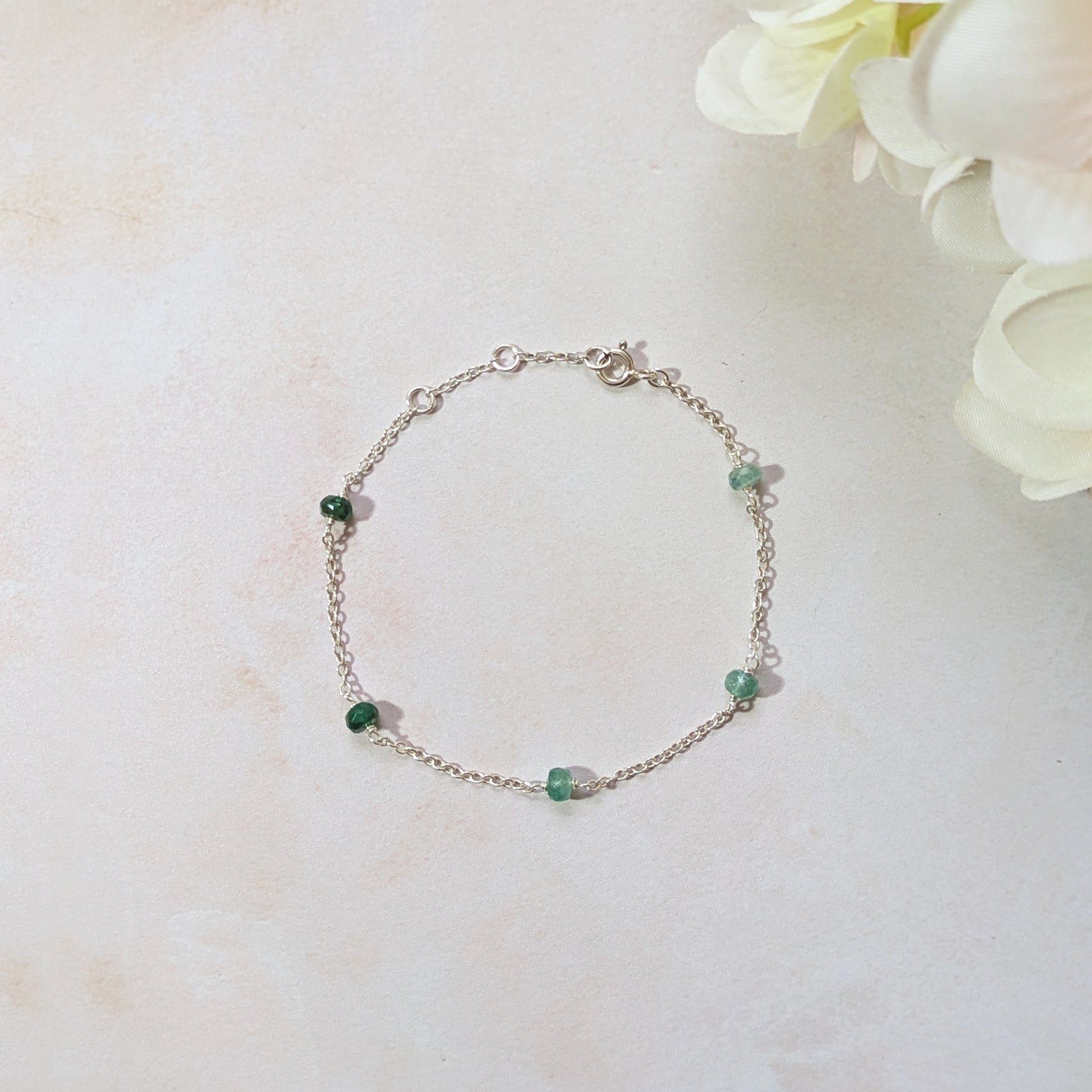 Silver chain bracelet with Emerald gemstone beads on a light surface with flowers.