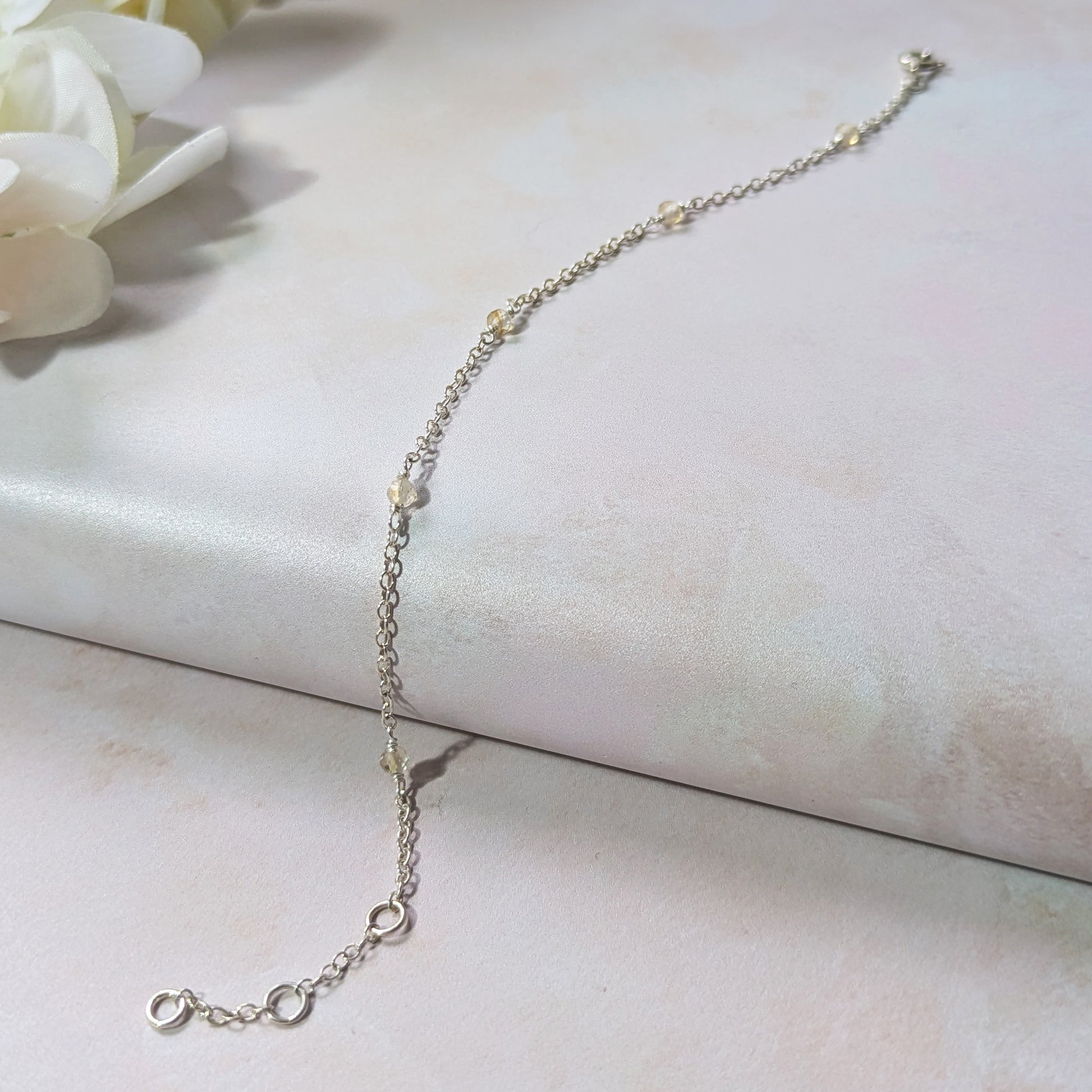 Silver stationed chain bracelet with Citrine gemstone beads on a light pink surface with white flowers in the corner.