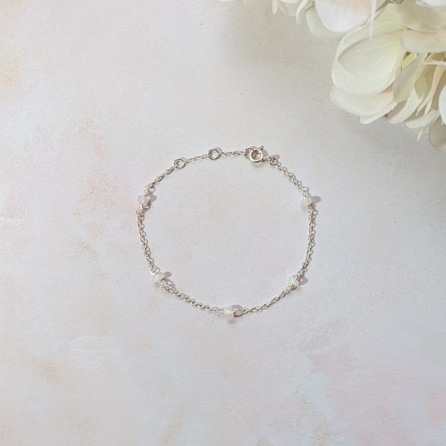 Silver stationed bracelet with small Opal gemstone beads on a light marble surface with flowers.