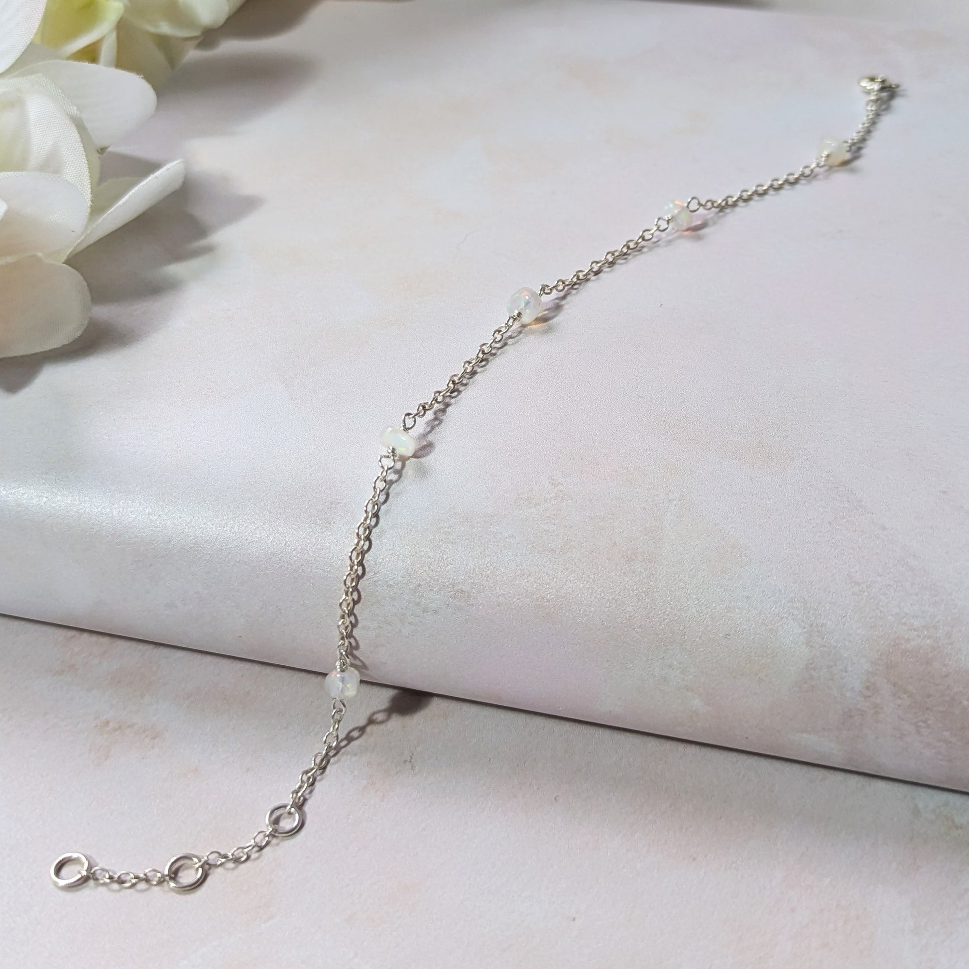 Silver stationed chain bracelet with Opal gemstone beads on a light pink surface with white flowers.