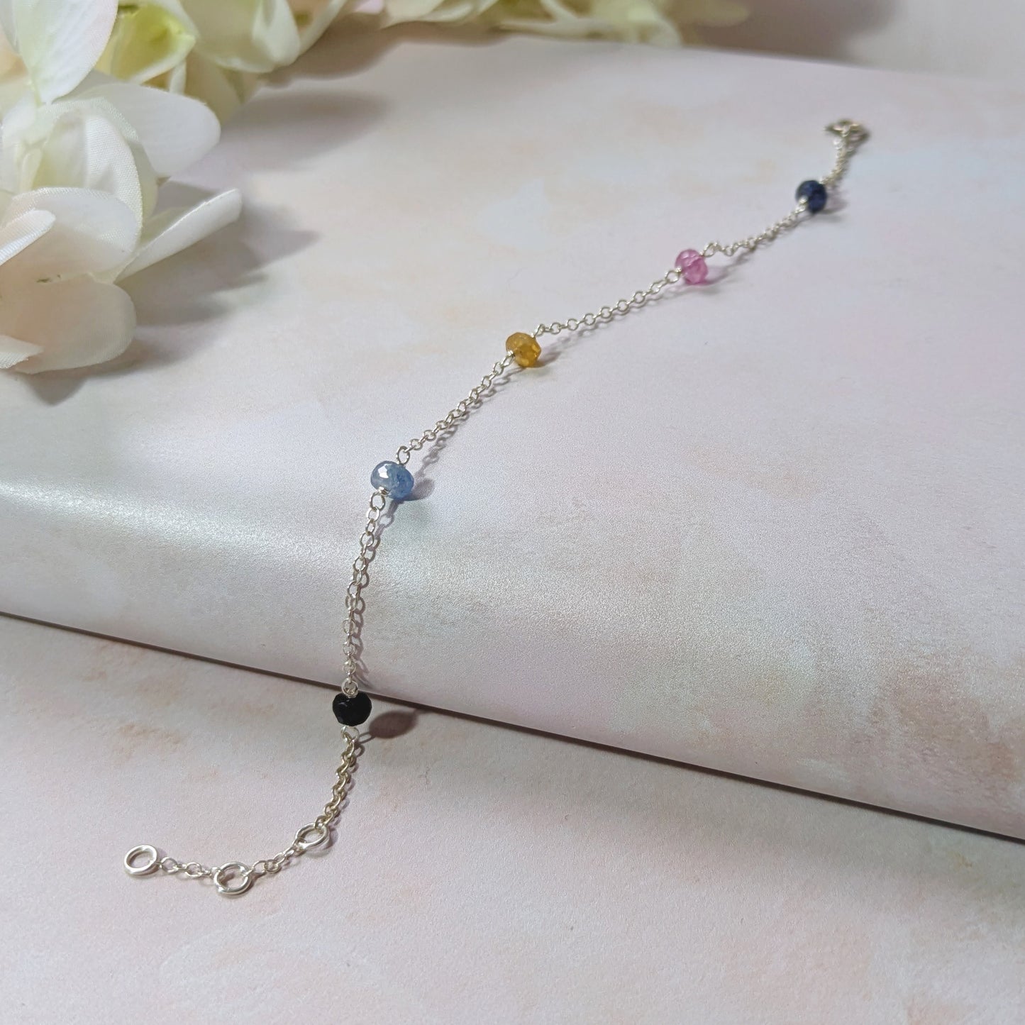 Silver stationed chain bracelet with multicoloured Sapphire gemstone  beads on a white surface with flowers in the background.