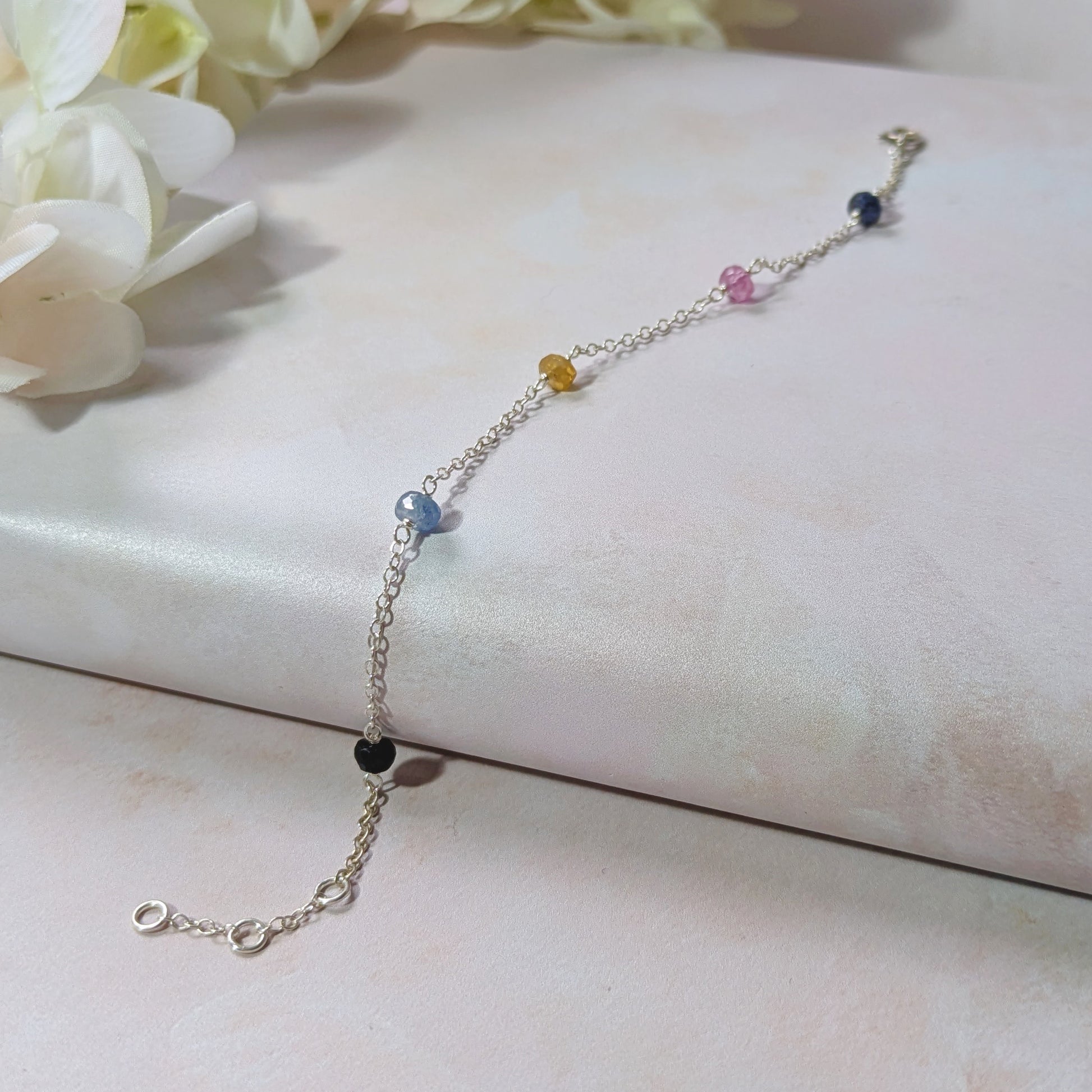 Silver stationed chain bracelet with multicoloured Sapphire gemstone  beads on a white surface with flowers in the background.