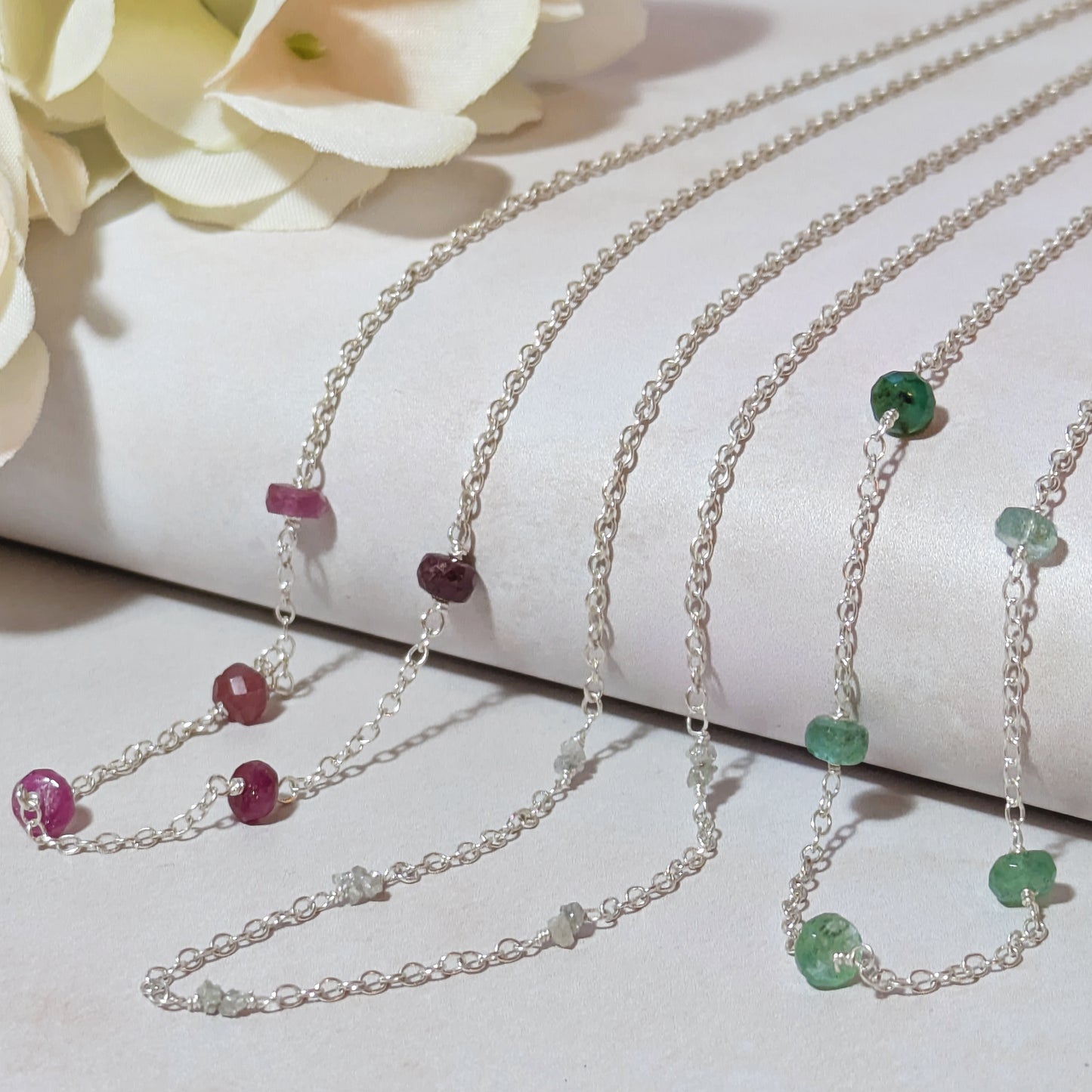 Three silver necklaces with Ruby, Emerals and rough Diamond nugget gemstone beads on a white surface with flowers in the background.