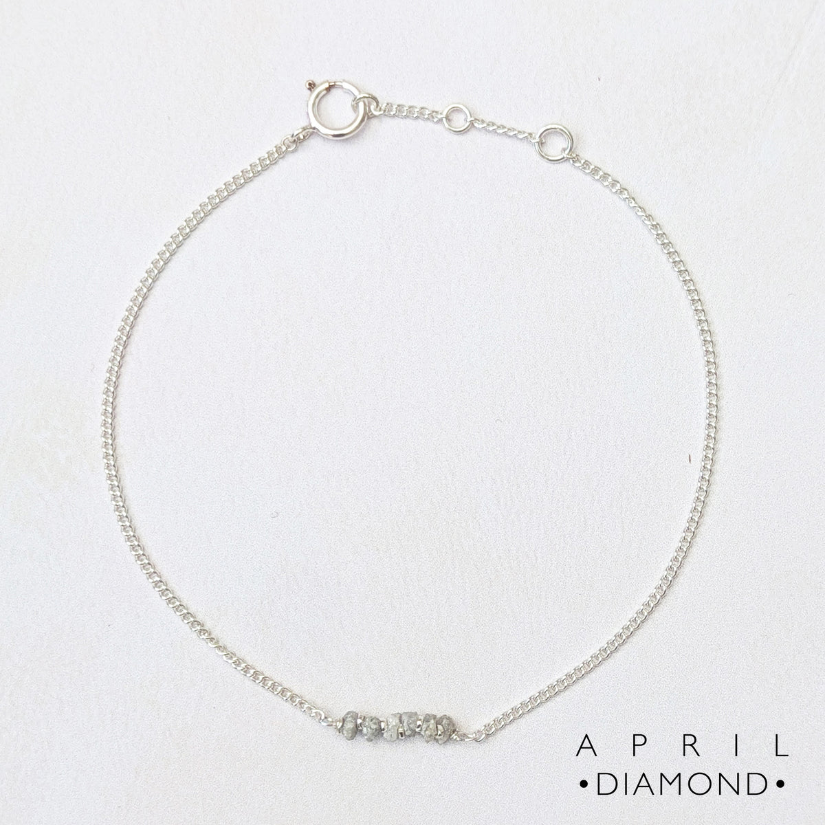 A delicate silver chain bracelet with a central bar featuring raw diamond nugget beads, displayed on a textured white background with the text 'APRIL DIAMOND' in the bottom right corner.