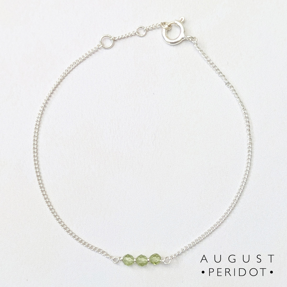 A delicate silver chain bracelet with three small light green peridot beads at the center, displayed on a clean white background with the text 'AUGUST PERIDOT' in the bottom right corner.