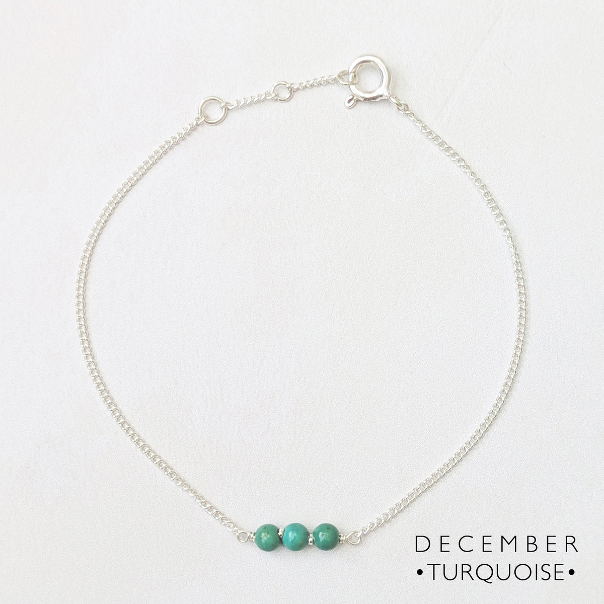 A delicate silver chain bar bracelet featuring three small turquoise beads at the centre, displayed on a clean white background with the text 'DECEMBER • TURQUOISE •' in the bottom right corner.