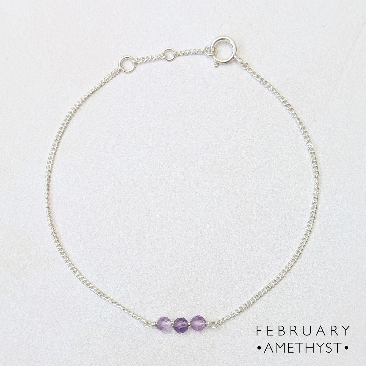A delicate silver birthstone bar bracelet with three small purple amethyst beads at the front, displayed on a textured white background with the text 'FEBRUARY • AMETHYST •' in the bottom right corner.