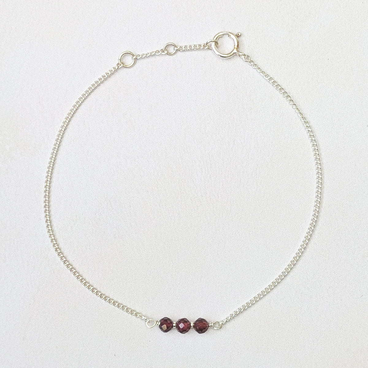 A delicate silver chain bracelet with three small, faceted dark red beads aligned horizontally at the centre, displayed on a light off-white textured background.