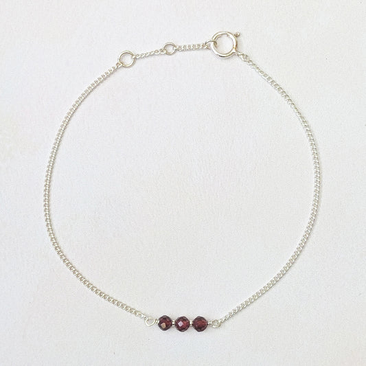 A delicate silver chain bracelet with three small, faceted dark red beads aligned horizontally at the centre, displayed on a light off-white textured background.