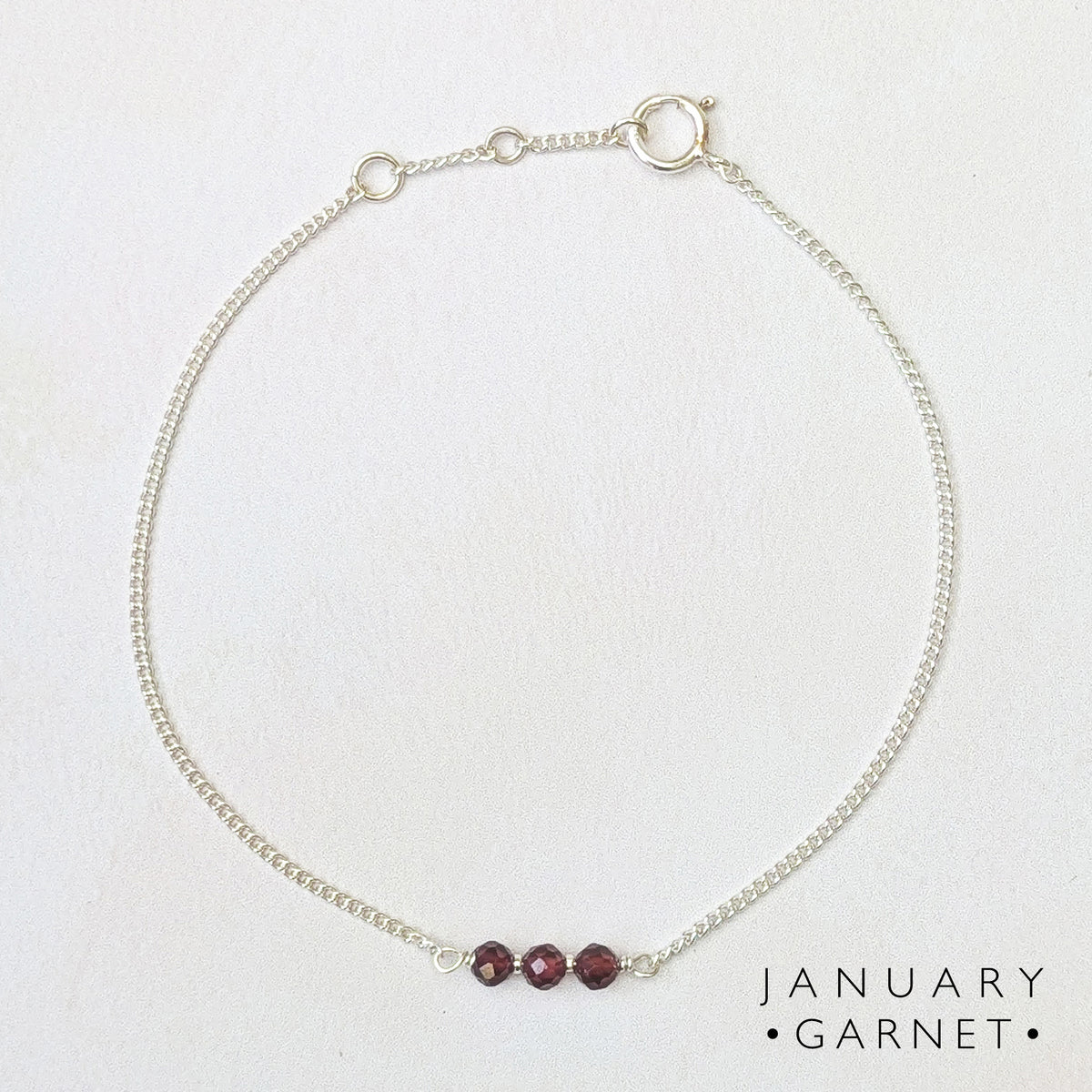 A delicate silver birthstone bar bracelet with three small dark red garnet beads at the front, displayed on a textured off-white background. The text 'JANUARY GARNET' is printed in the bottom right corner.