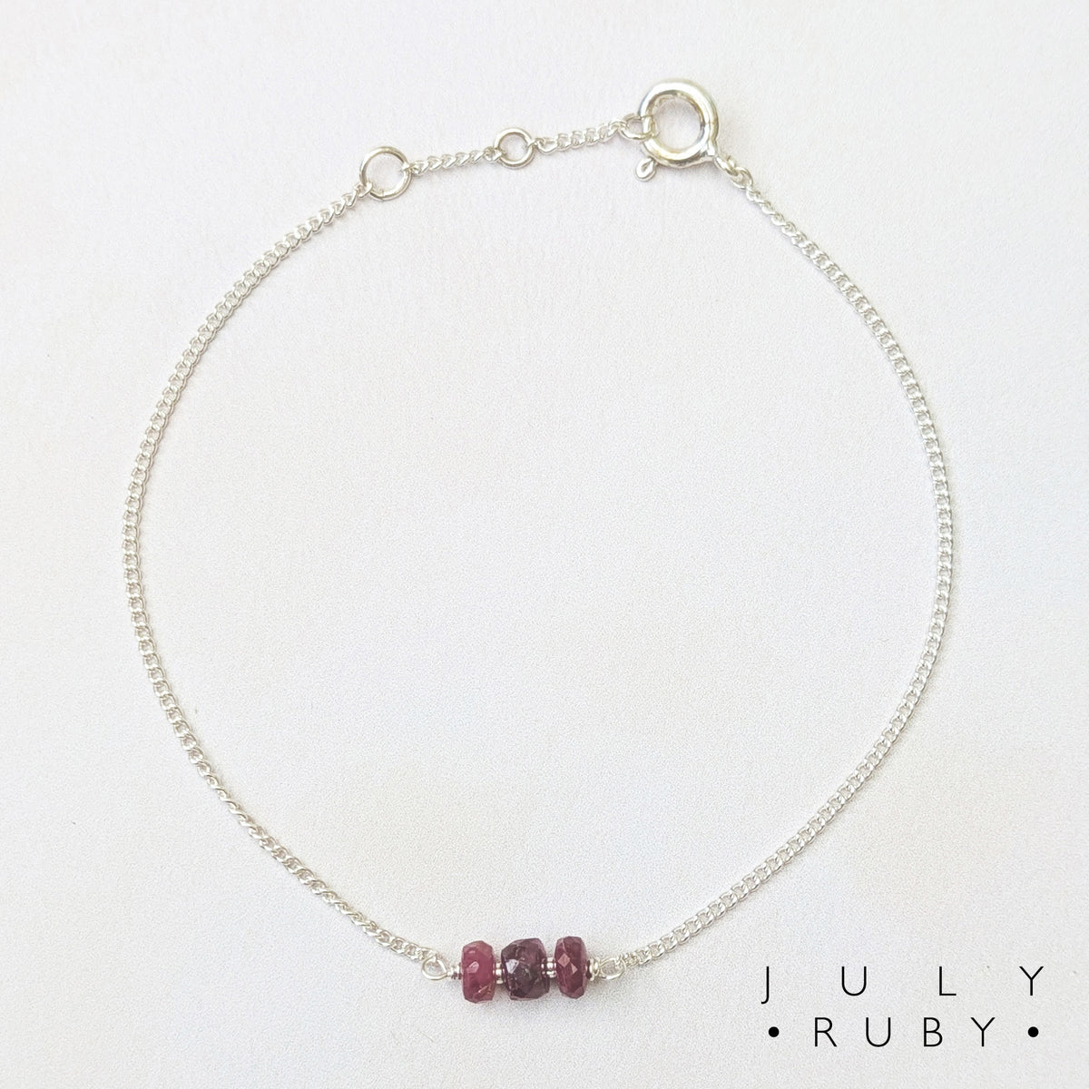 A delicate silver chain birthstone bracelet with three small, faceted dark red ruby beads at the centre, displayed on a clean white background with the text 'JULY RUBY' printed in the bottom right corner.