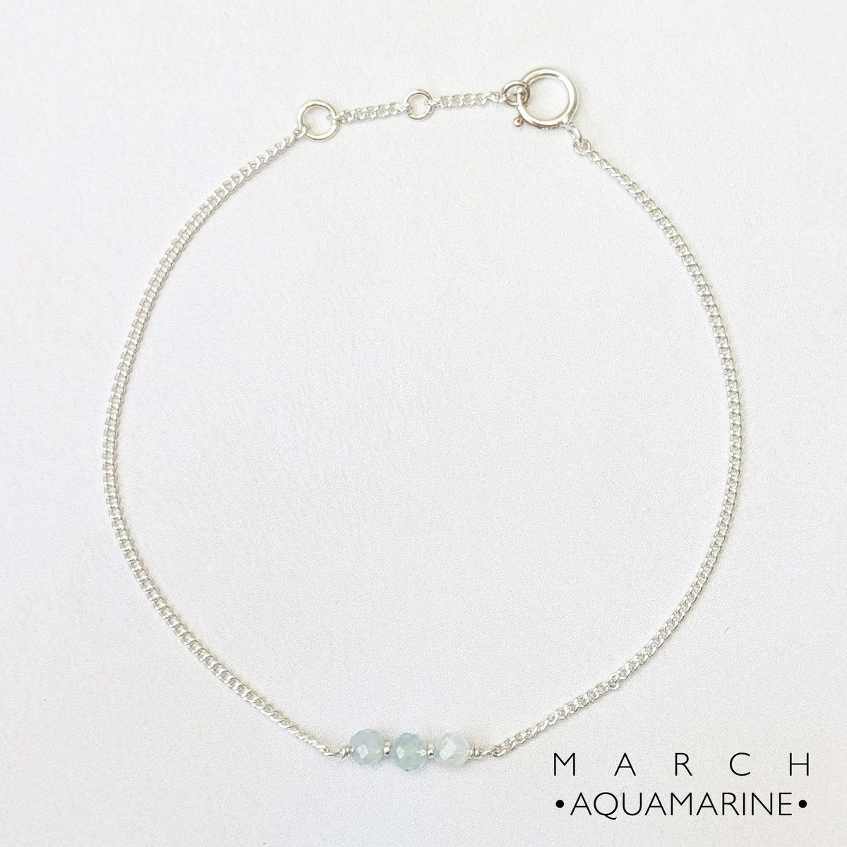 A delicate silver in line bracelet featuring three small aquamarine beads at the front, displayed on a clean white background with 'MARCH • AQUAMARINE •' text in the lower right corner.