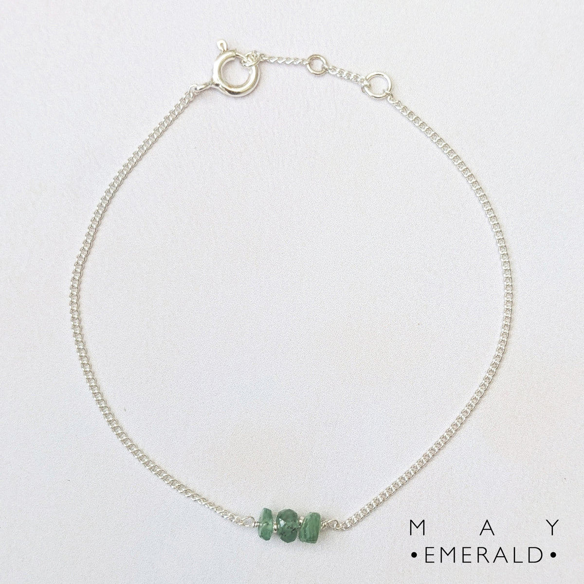 A delicate silver birthstone bar bracelet featuring three small emerald stones at the centre, displayed on a clean white background with the text 'MAY EMERALD' printed in the bottom right corner.