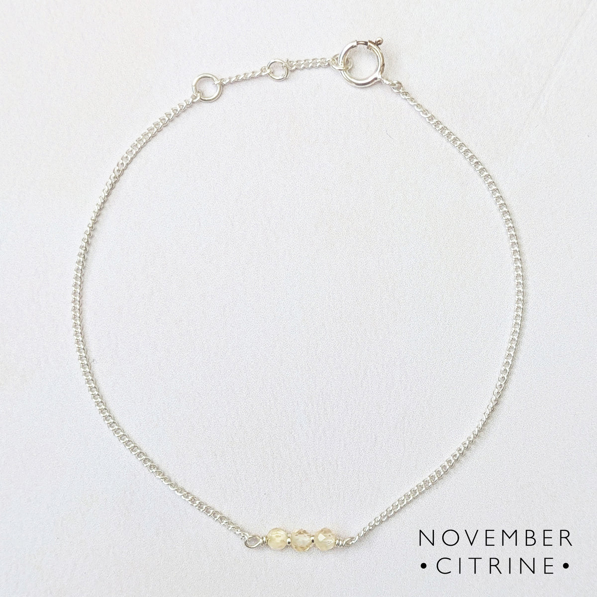 A delicate silver chain bracelet with three small pale yellow citrine beads at the front, displayed on a textured white background with the text 'NOVEMBER CITRINE' in the bottom right corner.