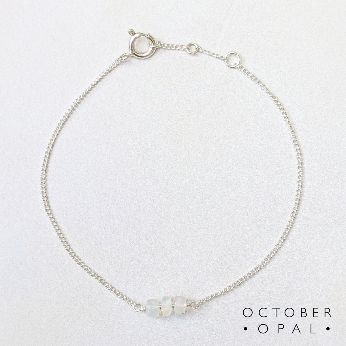 A delicate silver bar bracelet featuring three small, round, iridescent opal beads at the front, displayed on a clean white background with the text 'OCTOBER OPAL' printed in the bottom right corner.