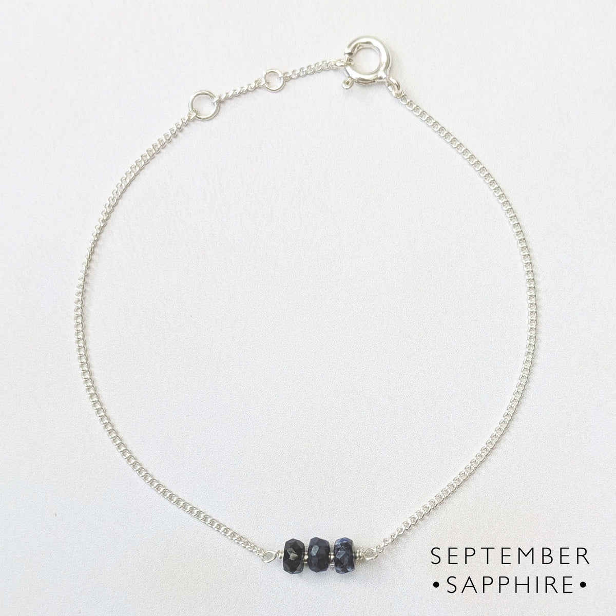 A delicate silver birthstone bar bracelet featuring three small, dark blue sapphires, displayed on a clean white background with 'SEPTEMBER SAPPHIRE' text in the lower right.