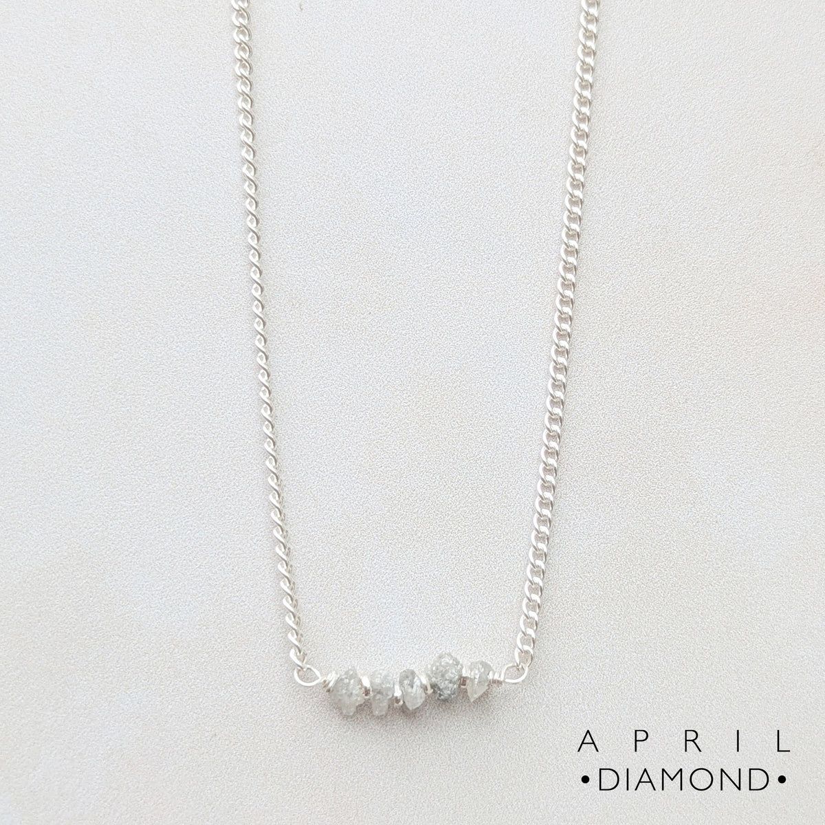 A delicate silver birthstone necklace featuring a horizontal bar with natural raw Diamond nugget beads, displayed on a clean white background with the brand name 'APRIL DIAMOND' in the bottom right corner.