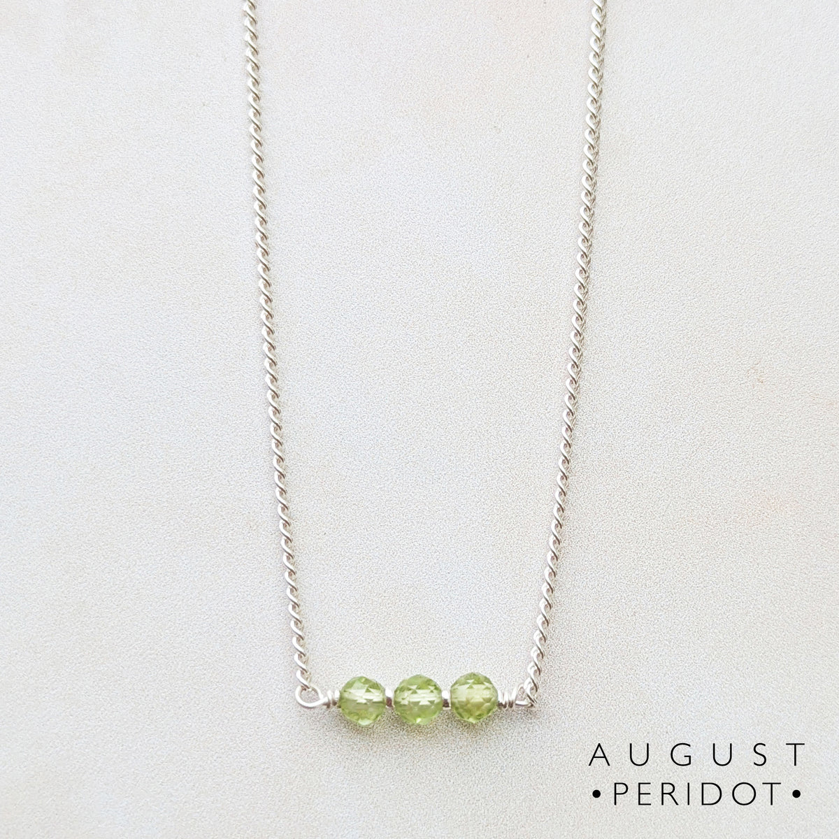 A delicate silver chain necklace featuring three small, faceted light green peridot beads aligned on a central bar, displayed on a clean white background with 'AUGUST PERIDOT' text in the lower right.