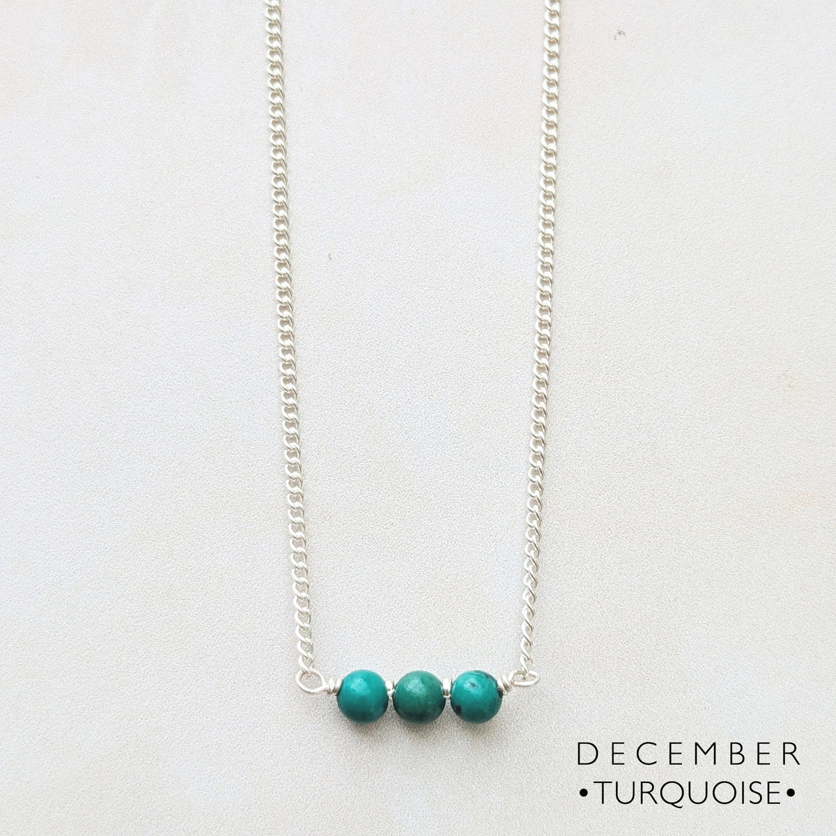 A delicate silver chain necklace featuring three small, round turquoise beads set on a bar against a plain white background, with the text 'DECEMBER • TURQUOISE •' printed in the bottom right corner.