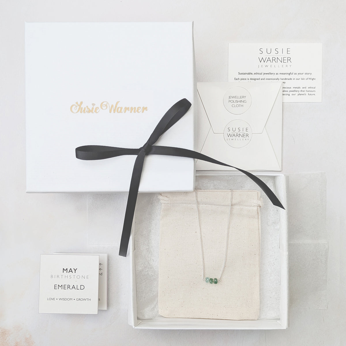 Susie Warner jewelry gift set featuring a delicate necklace with a small green stone pendant, presented in a white box with a black ribbon, alongside a white envelope, card, and gift box on a light marble background.