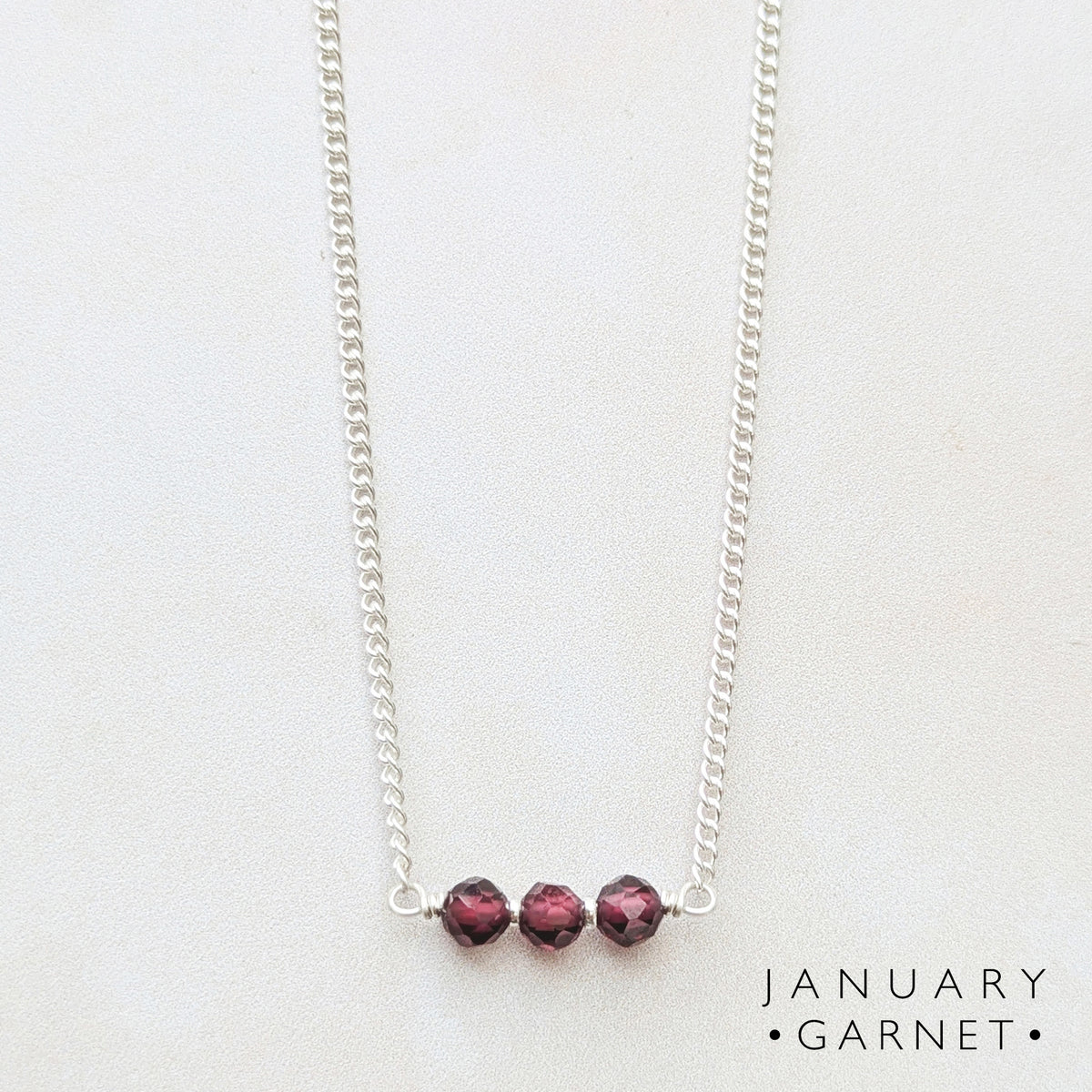 A silver birthstone bar necklace featuring three faceted dark red Garnet beads aligned horizontally, displayed on a light off-white background with the text 'JANUARY GARNET' in the bottom right corner.