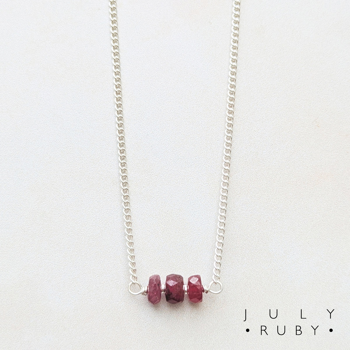 A delicate silver birthstone bar necklace featuring three small, faceted dark red ruby beads on a white background, with the text 'JULY RUBY' printed in the bottom right corner.
