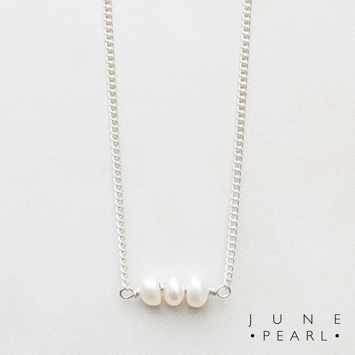 A delicate silver birthstone bar necklace featuring three small, lustrous white freshwater pearls aligned horizontally at the centre, displayed on a clean off-white background with the text  'JUNE PEARL' in the bottom right corner.