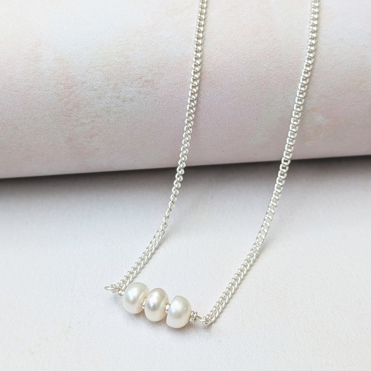 A delicate silver birthstone necklace featuring three small, lustrous white pearls aligned horizontally, displayed on a soft, light pink cylindrical stand against a pale background.