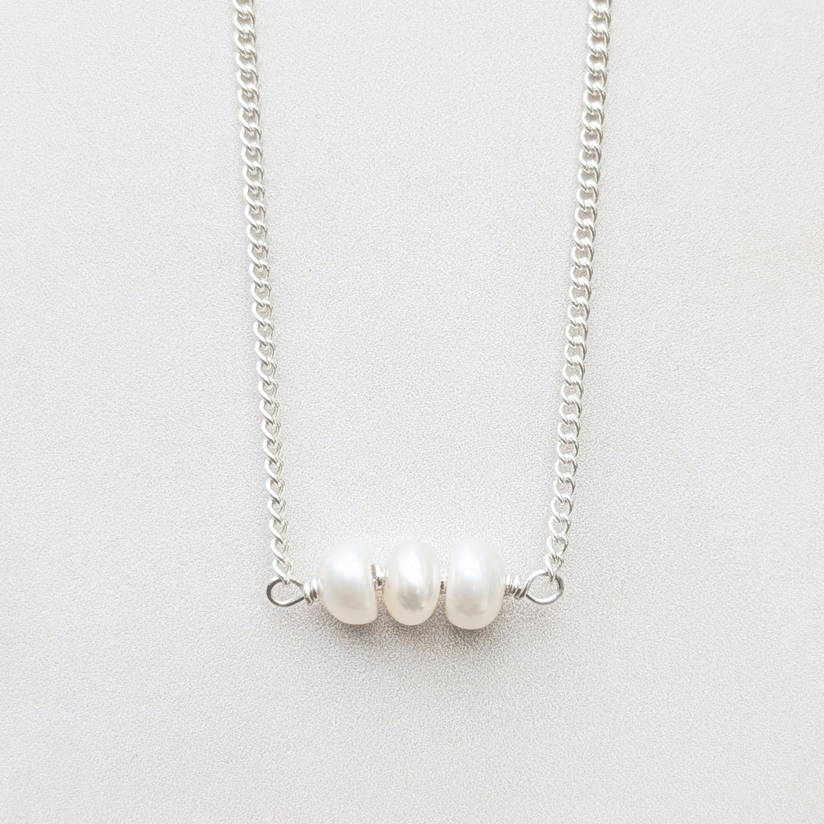 A silver birthstone bar necklace featuring three small, lustrous white pearl beads aligned horizontally at the centre, displayed against a light grey textured background.